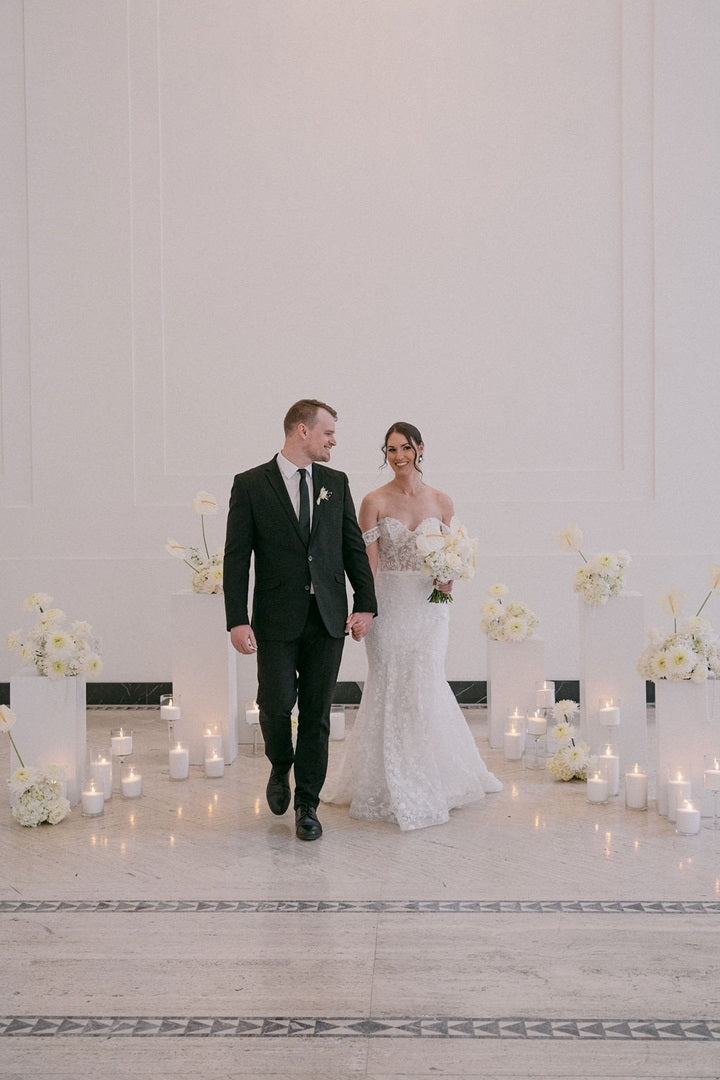 Bride in white gown walking with groom. She holds her bridal bouquet in one hand and is framed by floral arrangements in the background. Lit candles are placed in sets along the base of the pedestals.
