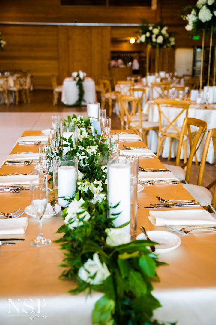 Table lined with white table setting, pillar candles, and loose greenery/florals lining the center of the table.