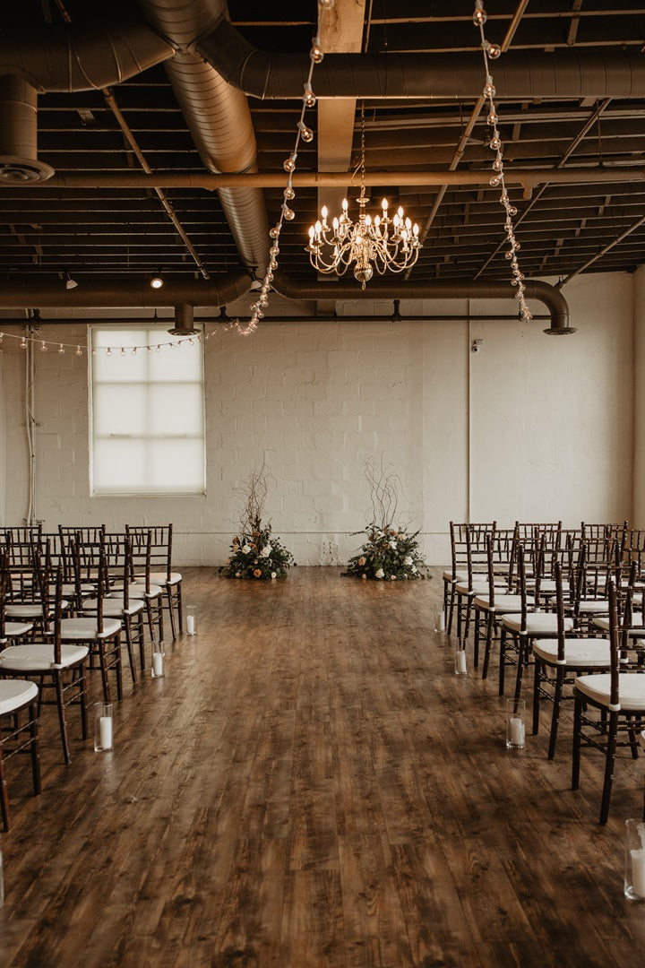 A center shot of the ceremony aisle. Chairs line either side, a chandelier hangs from the ceiling w/ string lights draping on along the rafters. Floral pieces lush with greenery and branches frame the end of the walkway creating an implied arch.
