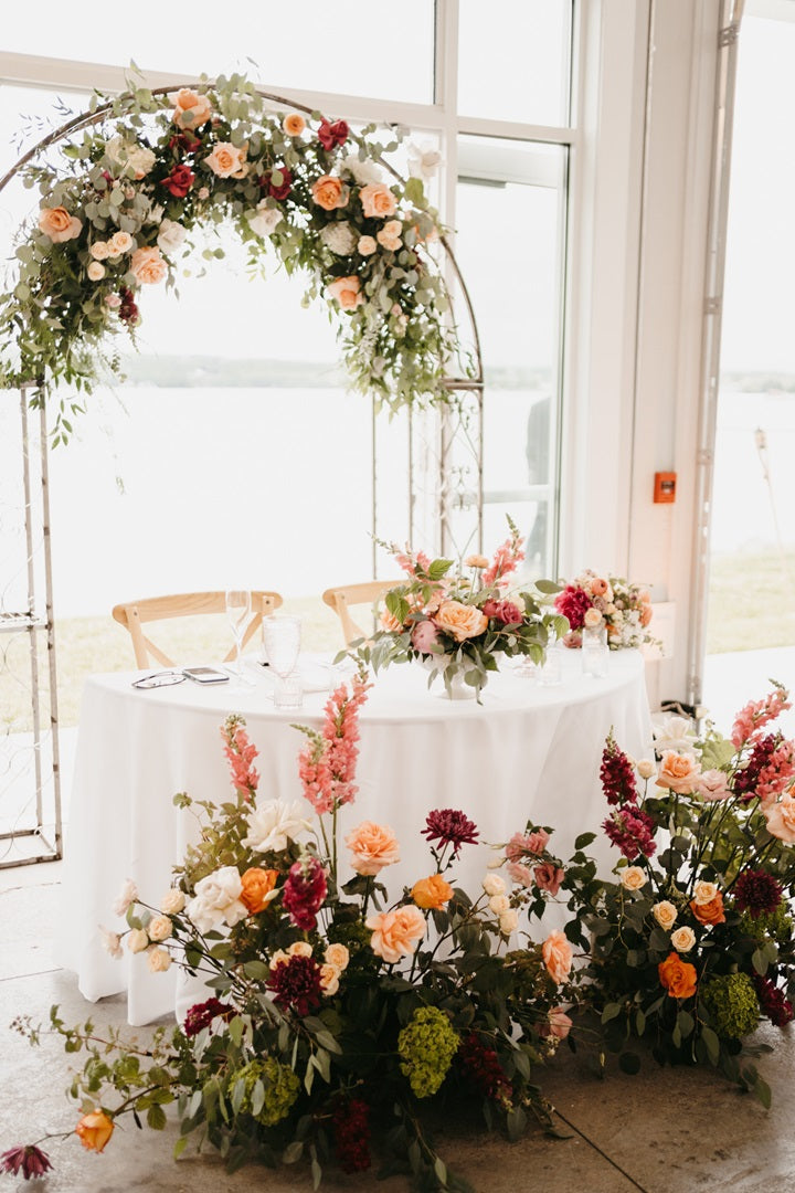 The sweetheart table with the floral arch, table centerpiece, and floor arrangements. All in the colorful palette with matching florals. Some florals shown are roses (traditional, garden, and spray), mums, hydrangea, snapdragons, and eucalyptus.