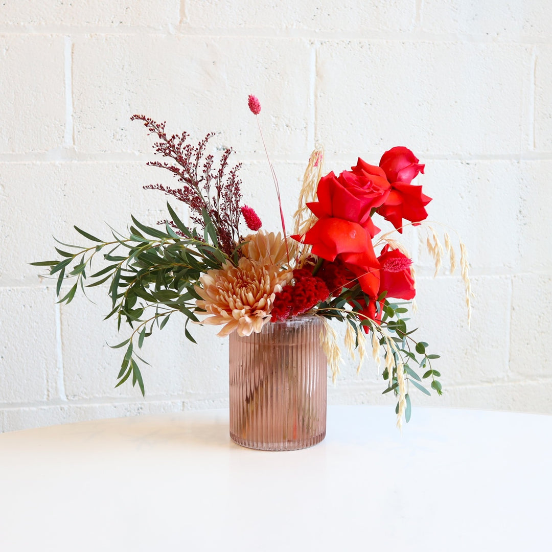 Brilliant | A vase arrangement with neutral mums, red roses, greenery, and red bunny tails in a transparent vase. Photo taken against a white background.