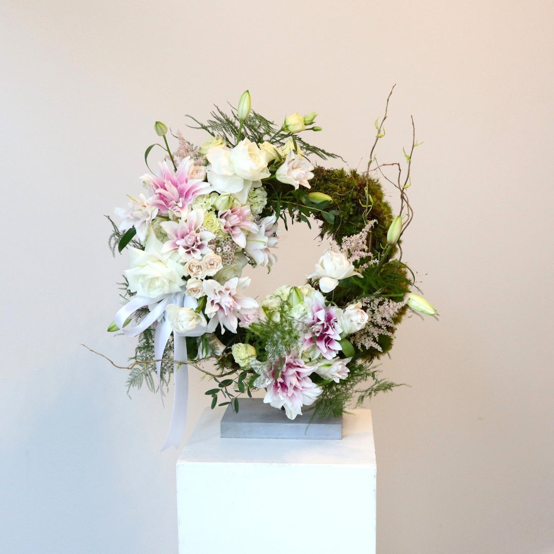 Garden Sympathy Wreath | A stunning wreath with an explosion of florals (roses, spray roses, double lilies, and more), decorative greenery, and natural moss. The colors are mostly white with accents of pink and green. Tied with a white ribbon and photo taken against a white background. Photo