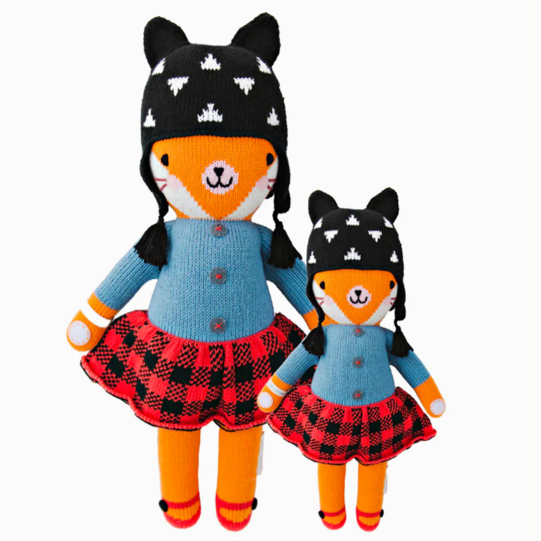 Sadie the Fox | Cuddle + Kind | A bright orange fox plush wearing a blue sweater, red plaid skirt, red shoes, and a black hat with tassels. 2 sizes shown, 20" and 13".