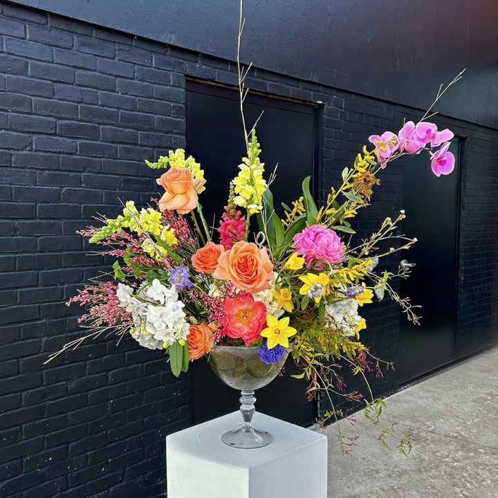 Stacy K Floral | A bright and natural flower arrangement with moss in a transparent glass compote. Some florals pictured are roses, snapdragons, hydrangea, orchids, and more.