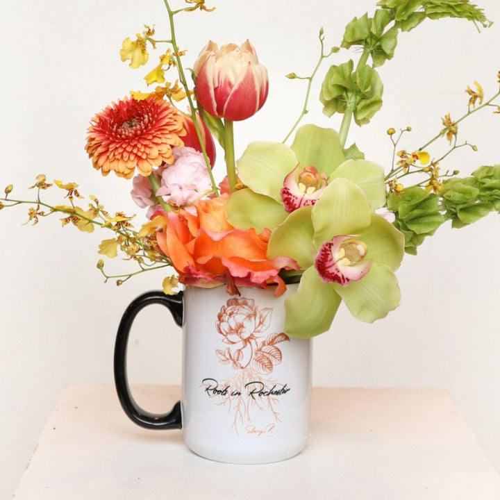 Stacy K Floral | Roots In Rochester Spring Mug | An orange and yellow arrangements in a white Roots in Rochester Mug. Some florals shown are roses, cymbidium orchids, oncidium orchids, double tulip, bells of ireland, and more.