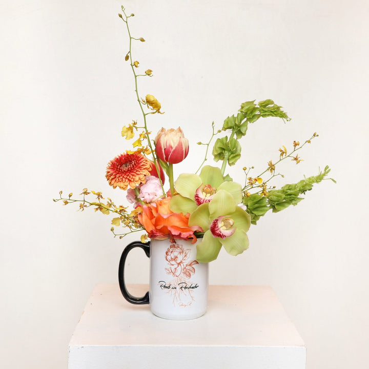 Stacy K Floral | Roots In Rochester Spring Mug | An orange and yellow arrangements in a white Roots in Rochester Mug. Some florals shown are roses, cymbidium orchids, oncidium orchids, double tulip, bells of ireland, and more.