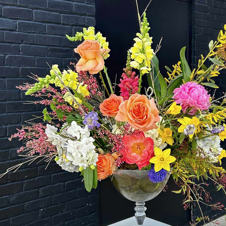 Stacy K Floral | A bright and rainbow colored floral arrangement with moss in a transparent compote. Some florals shown are roses, snapdragons, hydrangea, orchids, daffodils, and more.