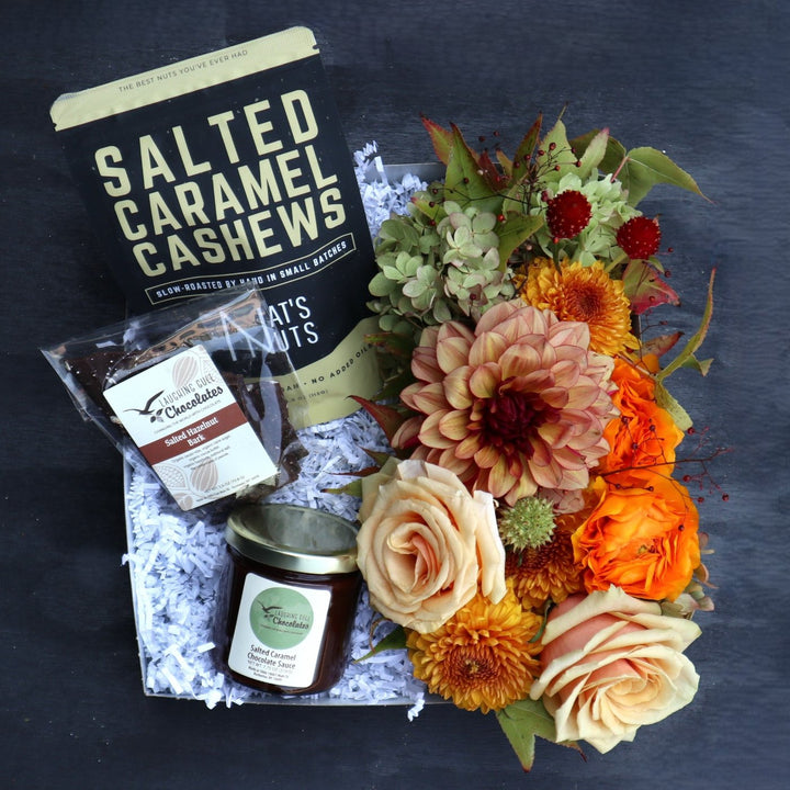 Gift box with salted caramel cashews, salted hazelnut chocolate bark, and a jar of salted caramel chocolate sauce, arranged next to a vibrant floral bouquet. The flowers include orange and peach roses, dahlias, and greenery, adding a warm, autumnal feel to the presentation. The box is lined with white crinkle paper for a polished look.