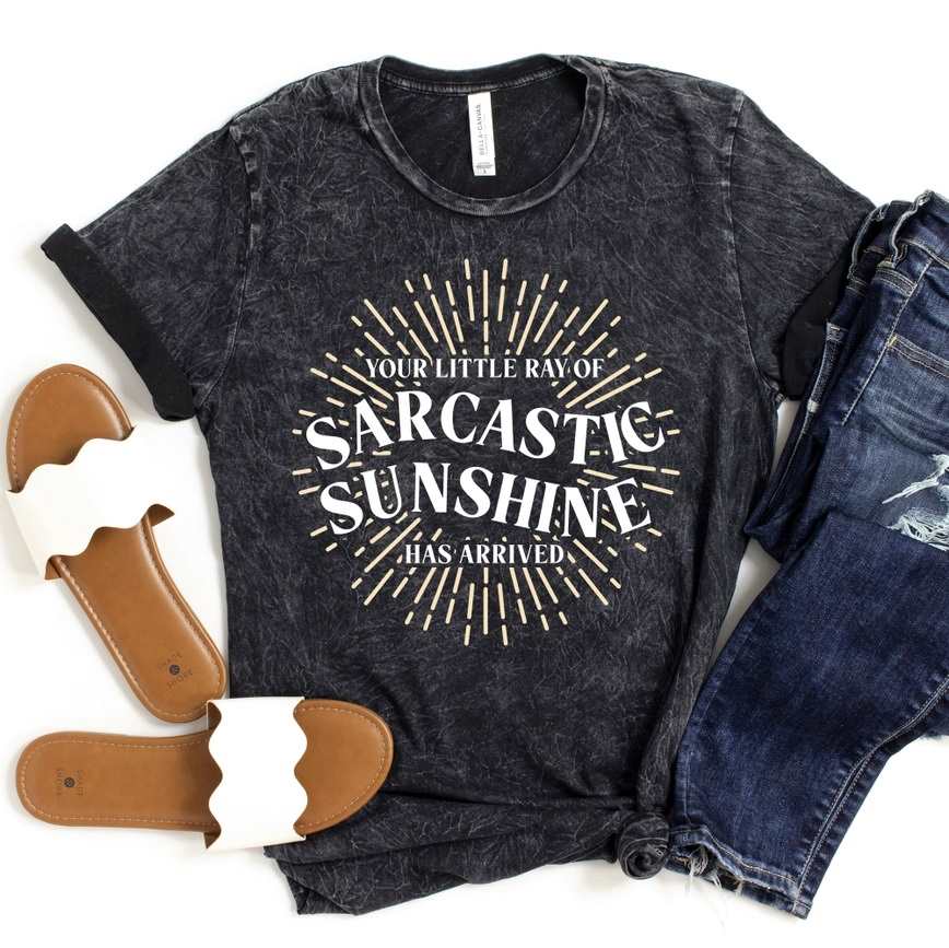 Mugsby | Your Little Ray of Sarcastic Sunshine Has Arrived | A washed black tshirt with text and yellow sunshine rays. White sandals and jeans accompany the shirt in the image.
