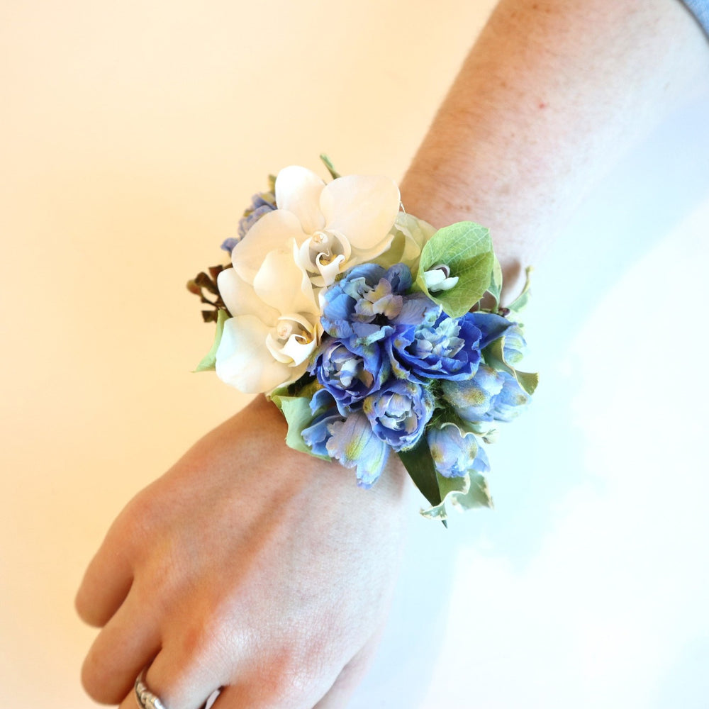 Corsage with two white mini orchids, blue delphinium, bits of bells of ireland with someone wearing it.