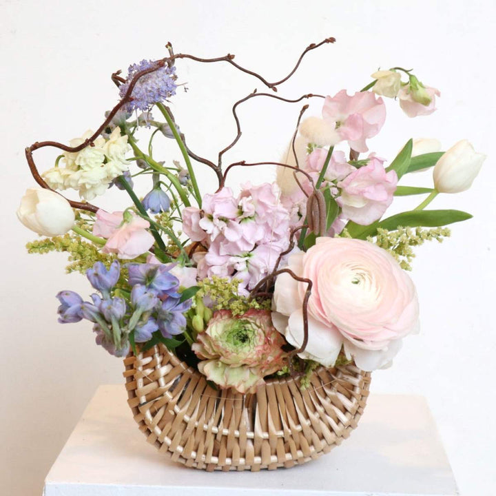Pastel spring flower arrangement with ranunculus, tulips, sweet peas, and hydrangeas in a woven basket with decorative branches.