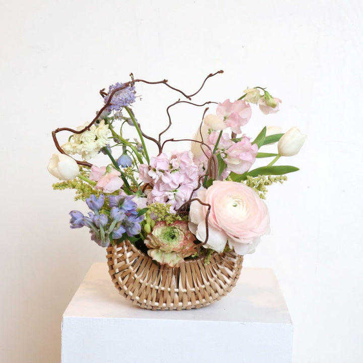 Pastel spring flower arrangement with ranunculus, tulips, sweet peas, and hydrangeas in a woven basket with decorative branches.