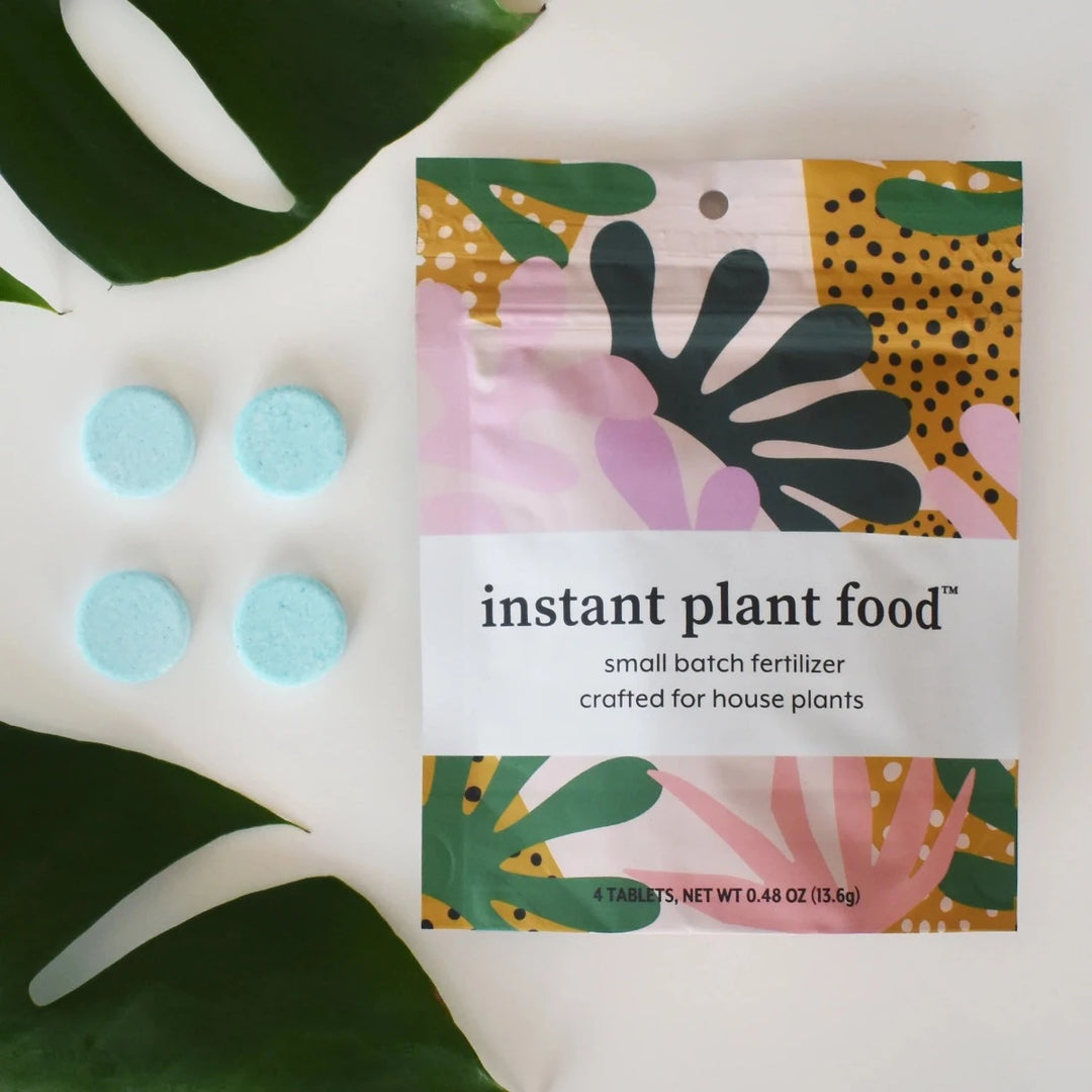 Instant Plant Food | A multi color packaged with abstract plants and text that reads "instant plant food, small batch fertilizer, crafted for houseplants, 4 tablets, net wt 0.48 oz, (13.6g)". Photo shows four blue tablets and the corner of monstera leaves as accents to the photo.