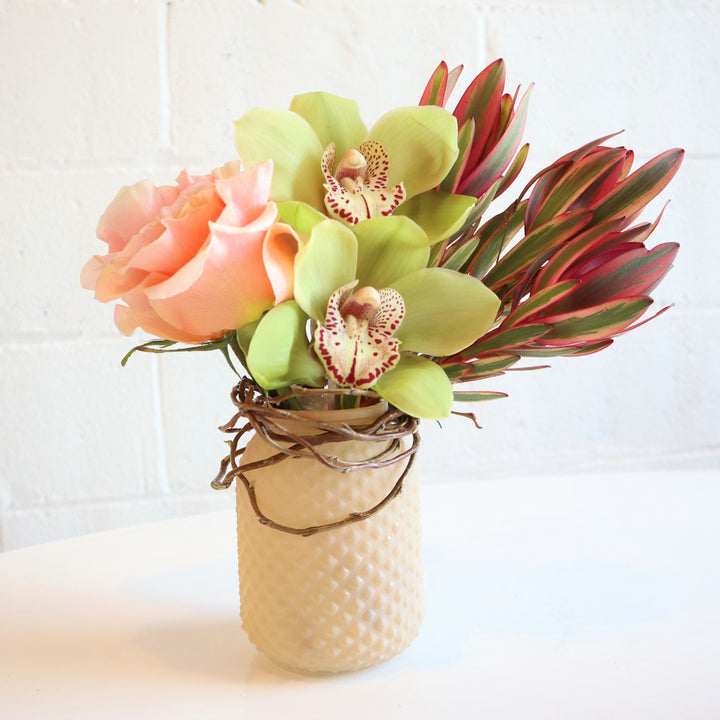 Glimmer | Close up on the vased arrangement with roses, orchid, and Leucadendron.  Accent branching is wrapped around the top of the vase. Colors are light pink and green.