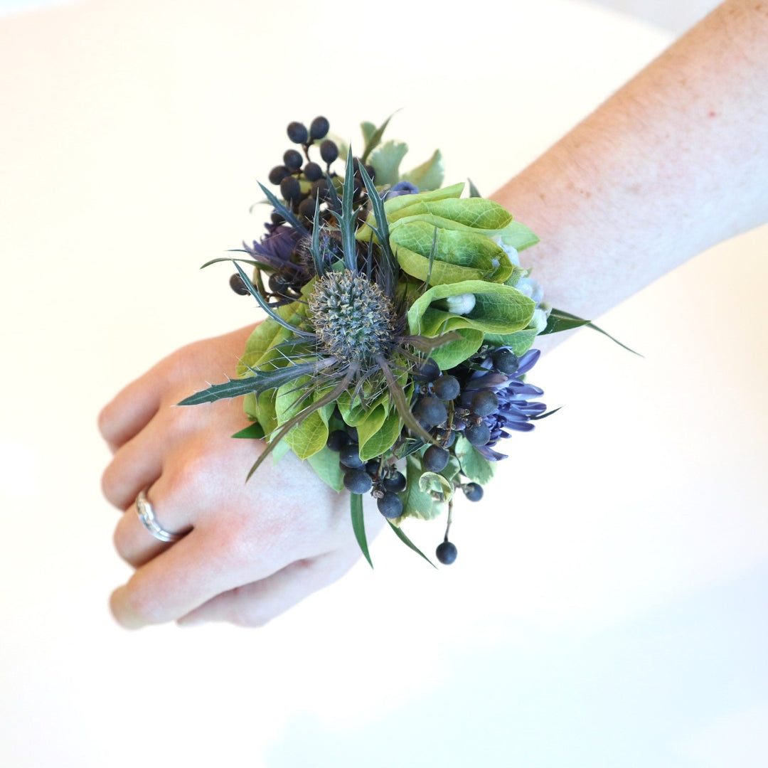 Arm wearing a corsage with bits of bells of ireland, blue thistle, blue berries and hints of blue mums.