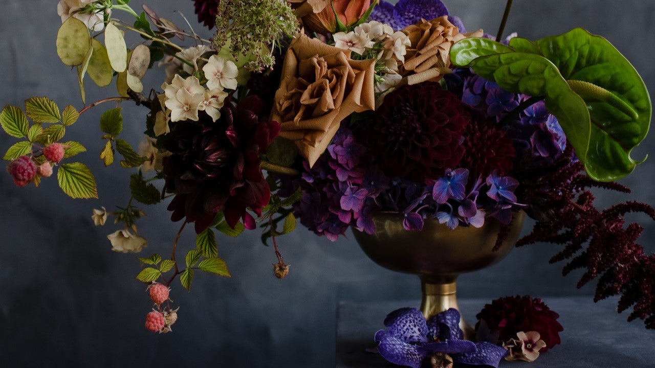 A dark and moody arrangement with a gold compote, hydrangea, raspberry plants, orchids, roses, and more.