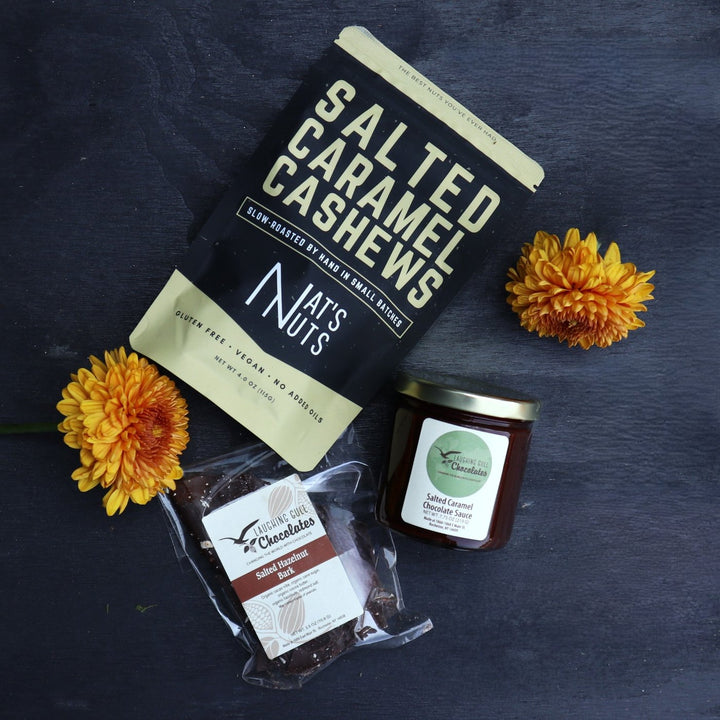 Bag of salted caramel cashews, a package of salted hazelnut chocolate bark, and a jar of salted caramel chocolate sauce arranged on a dark surface. Two orange chrysanthemums add a touch of color and complement the gourmet treats.