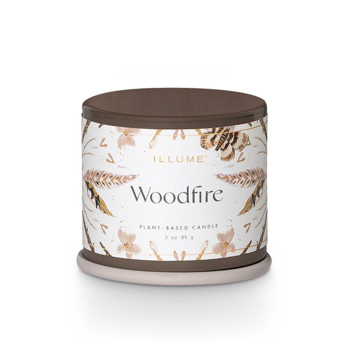Illume Demi Tin Candle | Woodfire | A brown tin candle with a white label decorated with moths, feathers, and maple seeds. Label reads "Plant based candle 3 oz, 85g."