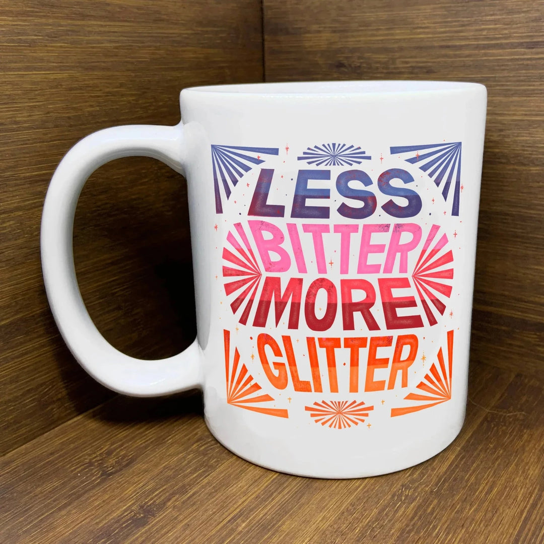Less Bitter More Glitter Mug | A white ceramic mug with less bitter more glitter written in purple, pink, red, and orange.