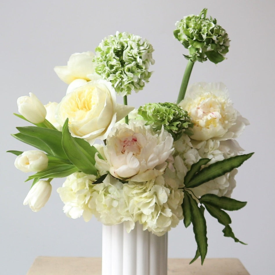 Limelight | A floral arrangement with whites, greens, and green blend florals. Arrangement is in a white vase on a cream pedestal against a white background. Close up shot of the arrangement.