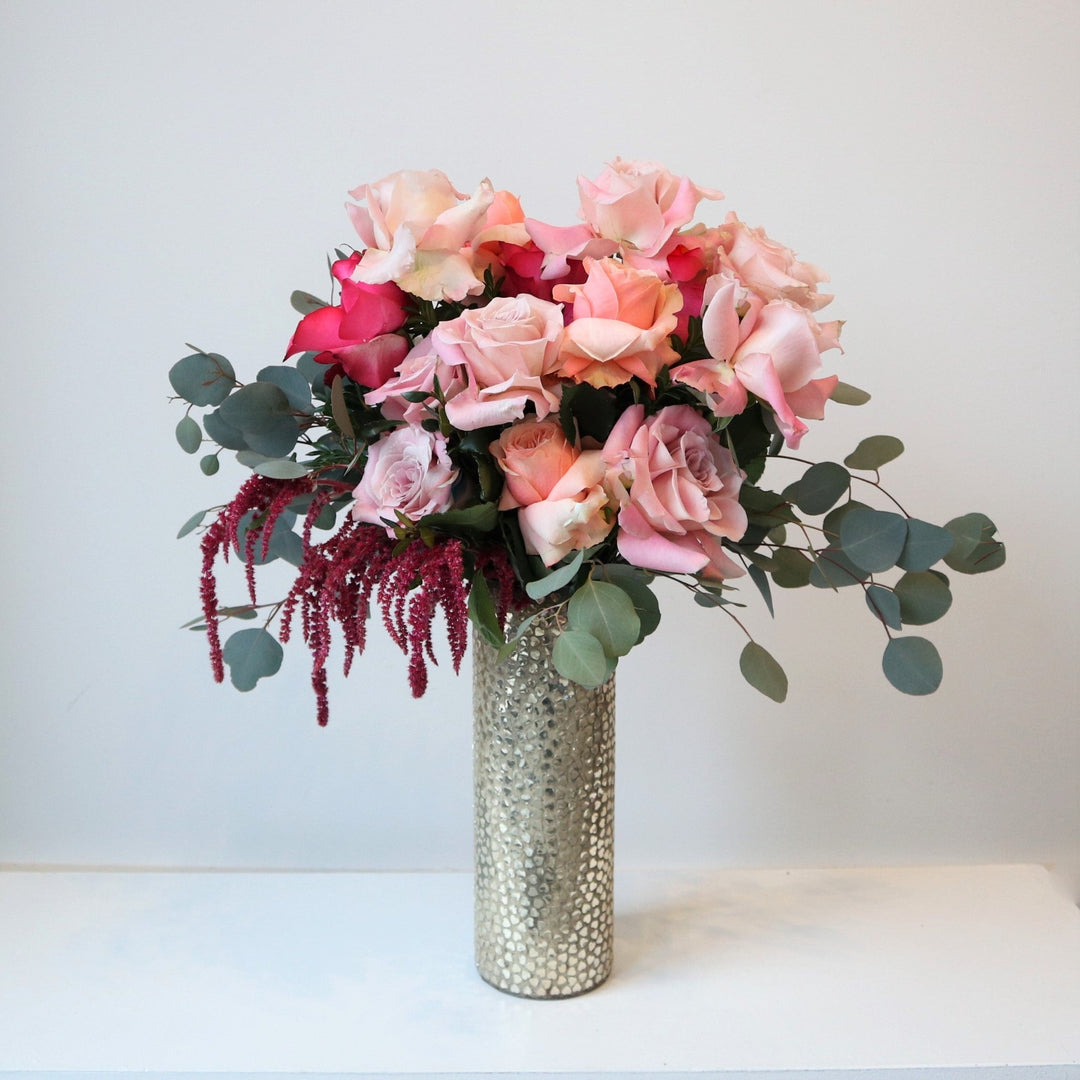 A pink and green floral arrangement in a tall, reflective vase. Showcases roses, eucalyptus, ameranthus, and other greens.