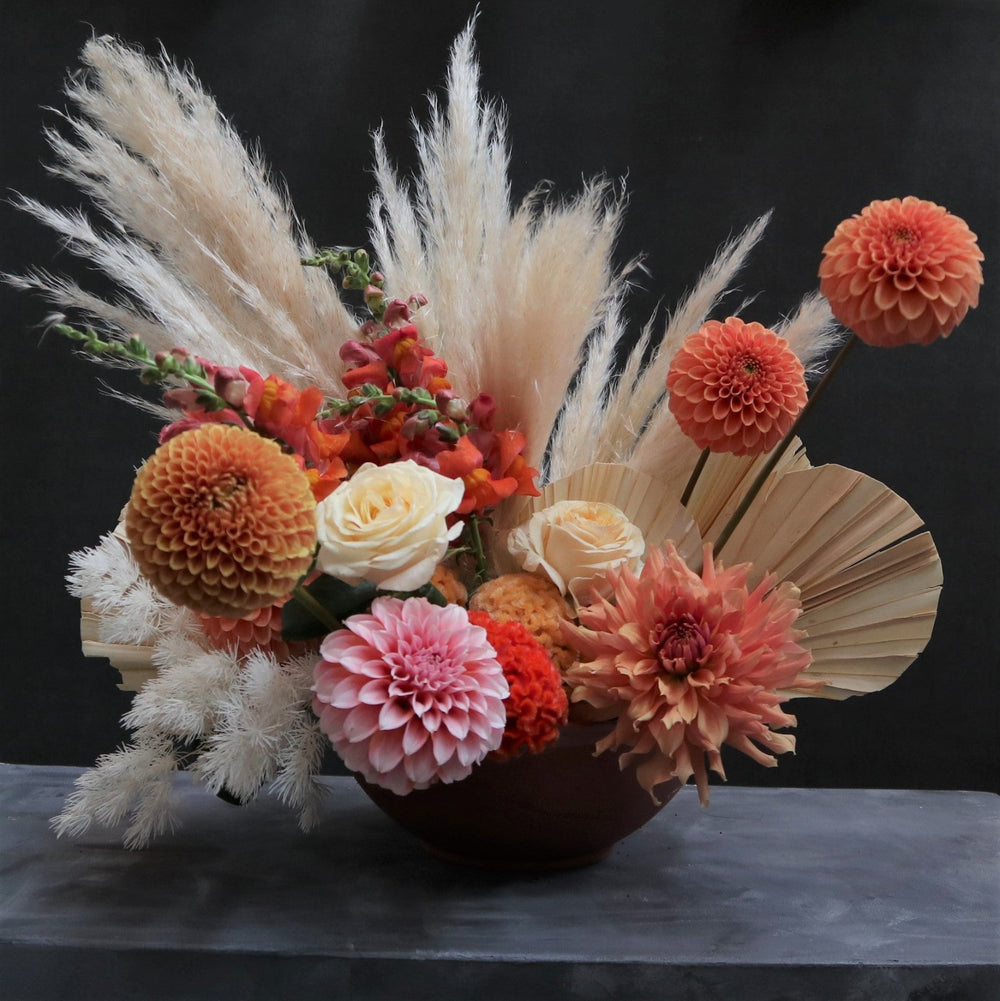 Orange vase, filled with orange dahlias, orange snapdragons, fan palms, cream pampas and celosia taken on a dark background.
