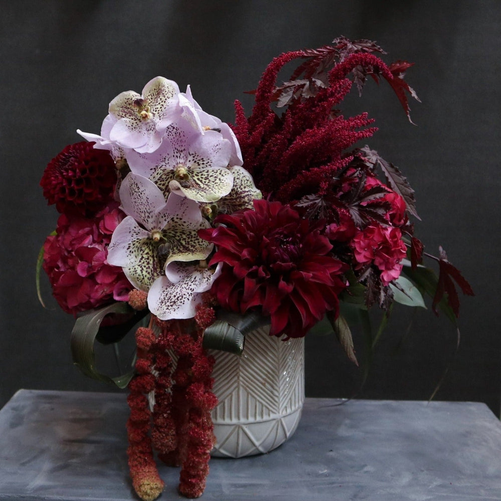 Cream vase with geometric pattern, filled withmaroon dahlia, pink hydrangea, red amarathus, maroon greenery with a large light purple orchid in the center. Placed on a dark background to show color variation.