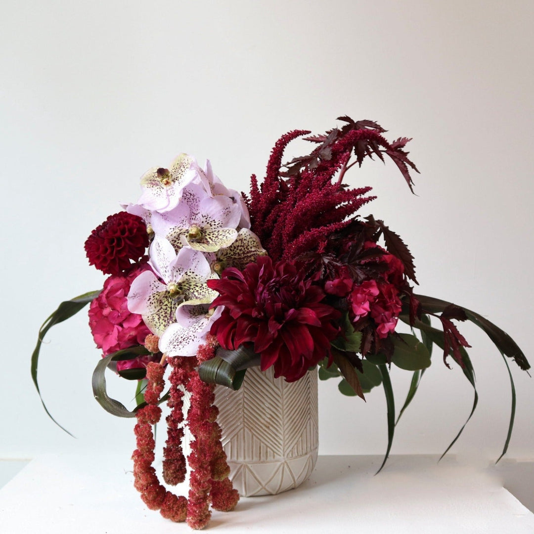 Cream vase with geometric pattern, filled withmaroon dahlia, pink hydrangea, red amarathus, maroon greenery with a large light purple orchid in the center.