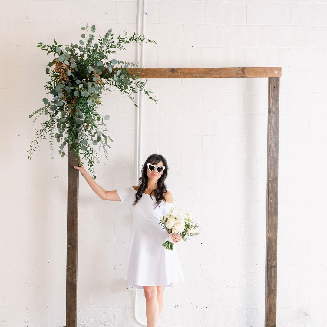Greenery Arch Piece | A greenery arch piece with eucalyptus and other greens. Attached to a rectangle natural wood arch. In the photo a model is posing in a white dress with white sunglasses and a bridal bouquet.