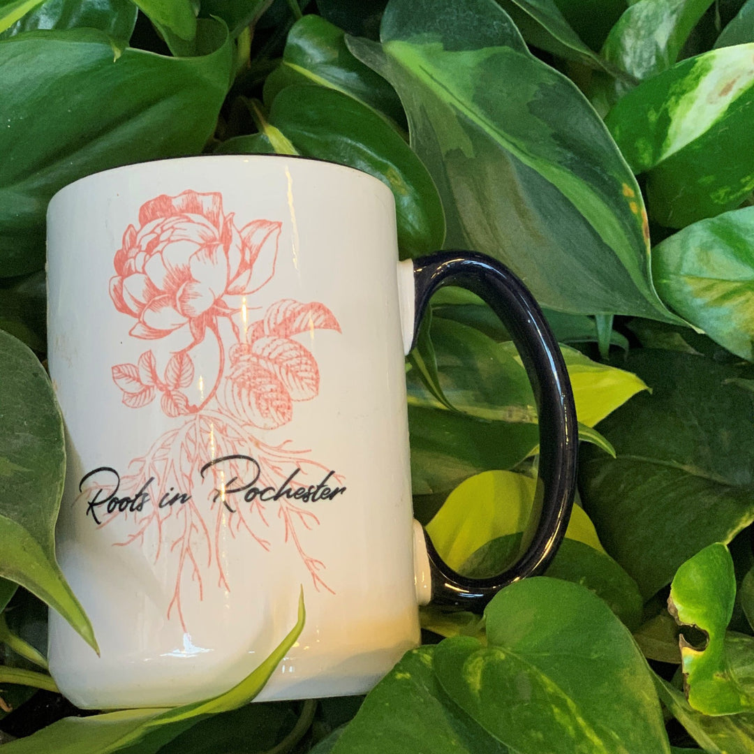 Roots in Rochester mug with pink floral design. Displayed against plant leaf background.
