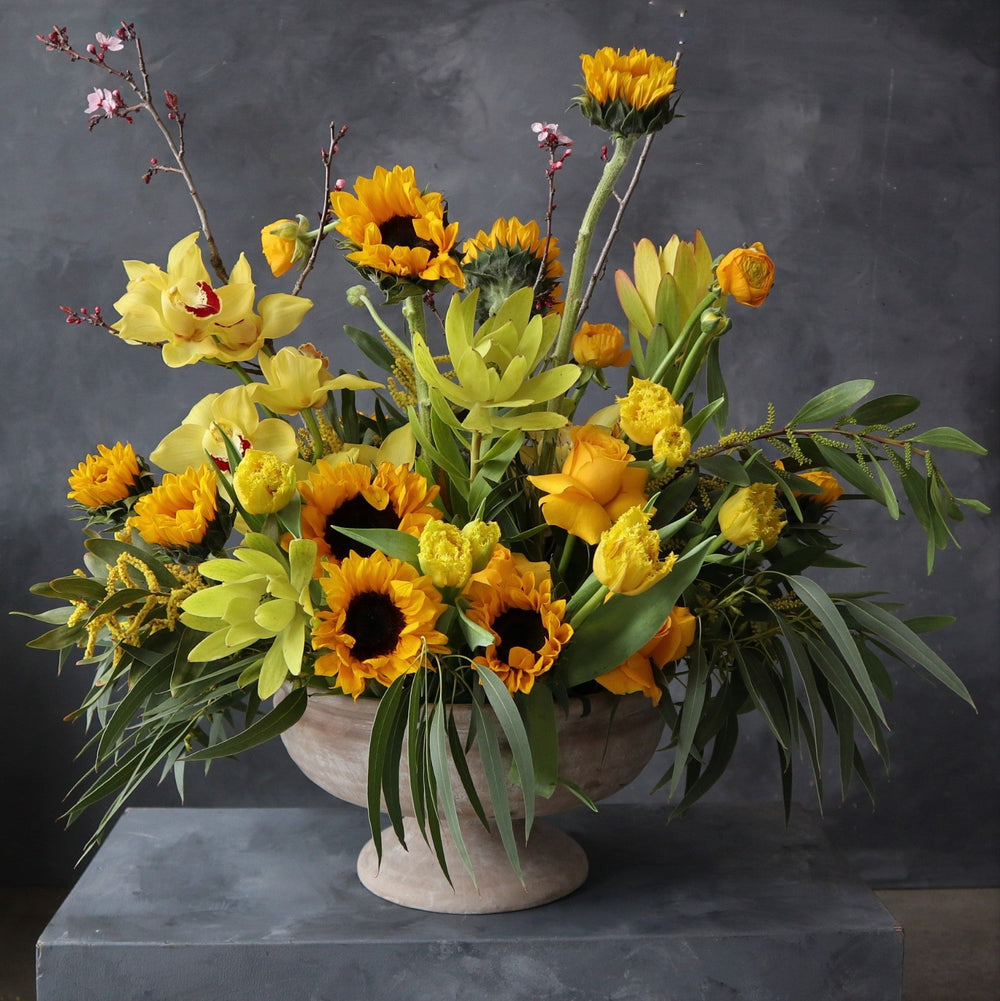 Yellow sunflowers, protea, orchids, roses and greenery.  Vased in a cream compote.