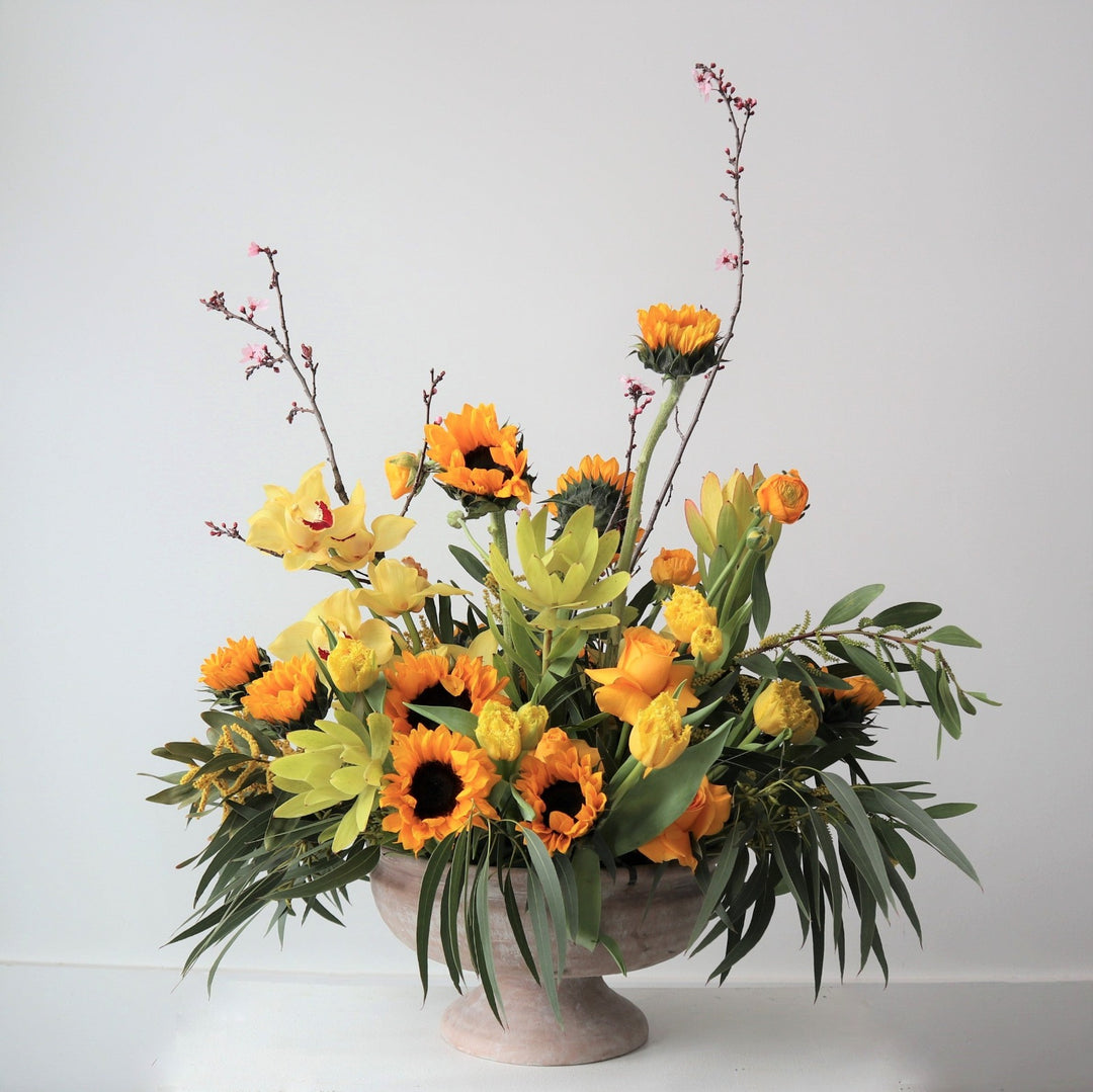 Yellow sunflowers, protea, orchids, roses and greenery.  Vased in a cream compote.