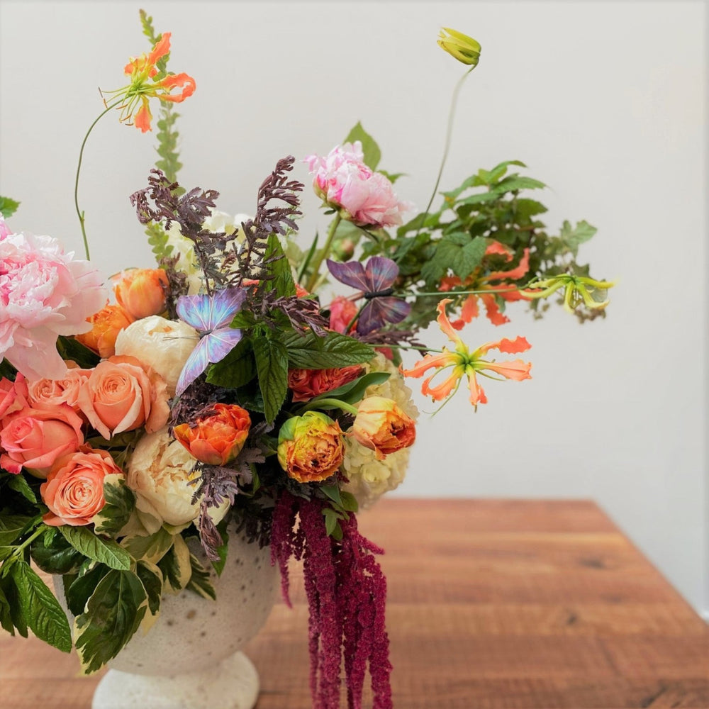 Large, organic style vased arrangement, with coral roses, pink peonies, butterflies, foliage and red hanging amaranthus, designed in a white vessel.