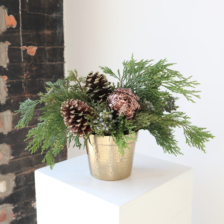 Arrangement in a gold metal vase, with evergreens, pinecones and an ornament.