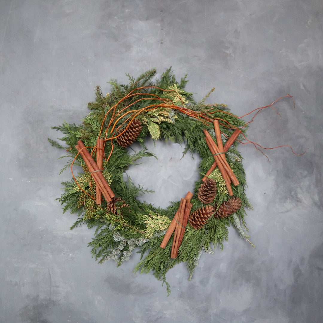 Evergreen wreath decorated with branching, different types of cedar, pinecones and cinnamon sticks. Photo taken on gray backdrop.