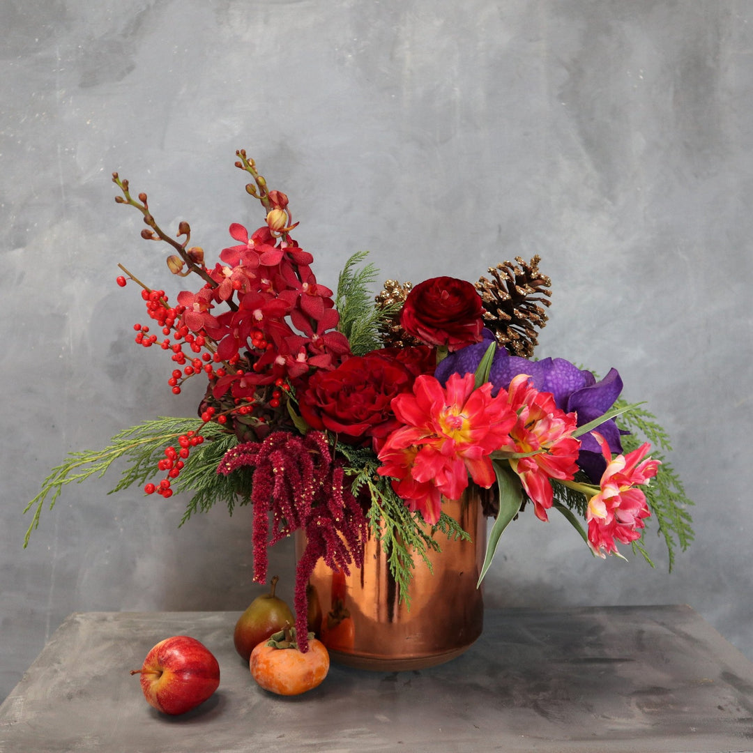Arrangement in a rose gold vessel, with pink tulips, red roses, red mokara roses, burgundy ranunculus, vanda orchids, red amarathus, pinecones, red ilex berries, wintergreens. Taken on a gray background. 