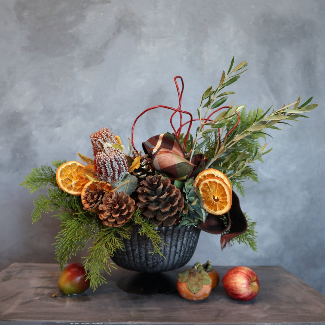 Arrangement in blue compote vessel, with pinecones, dried oranges, protea,evergreen, olive branches, curly willow and ribbon accents. Fruit at the base for styling purposes.
