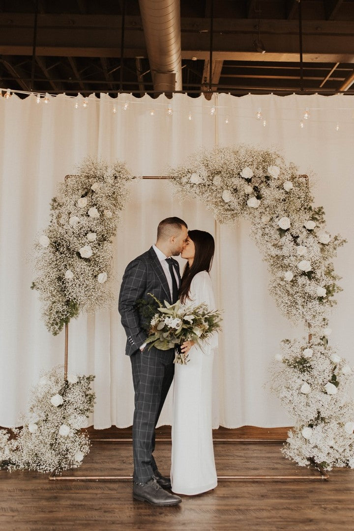 Bride and groom kiss in front of floral wedding arch at Arbor Loft. The arch is lush with roses, baby's breath, and greenery.