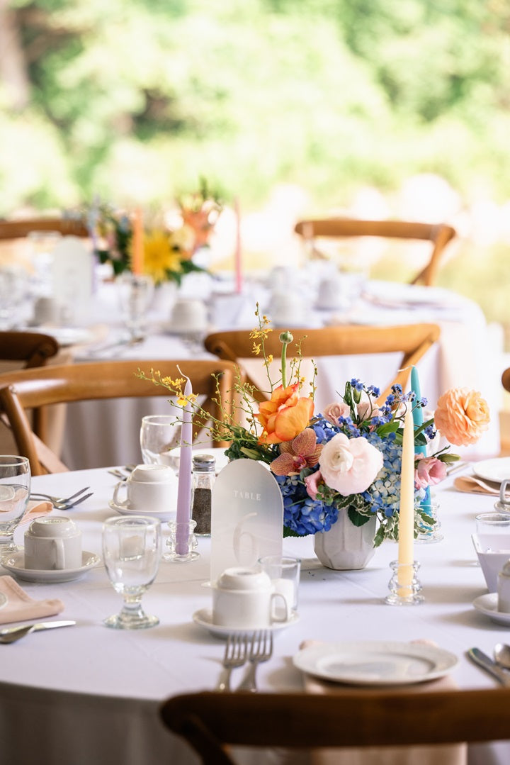 A table setting with a colorful floral centerpieces in orange, pink, blue, yellow, and green. Accent with purple, yellow, and blue, taper candles.