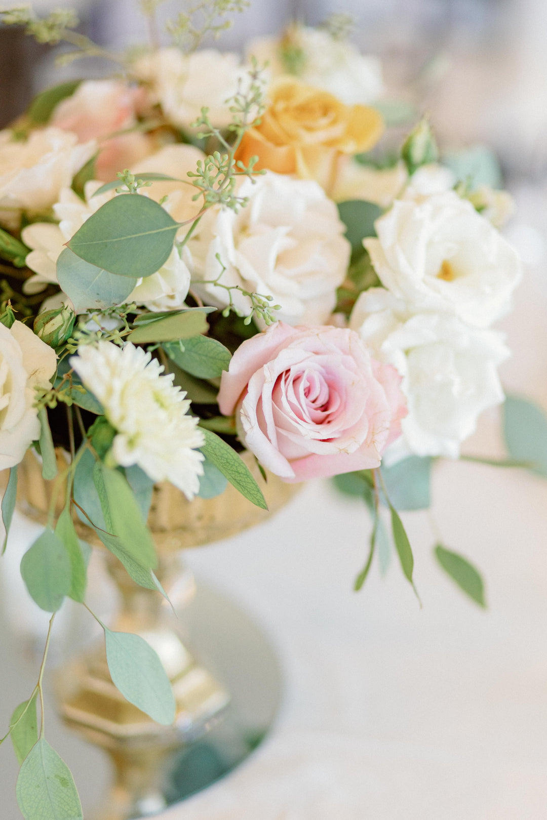 Close up on a floral centerpiece in a gold vase filled with pastel whites, pinks, and oranges. Accented with greenery.