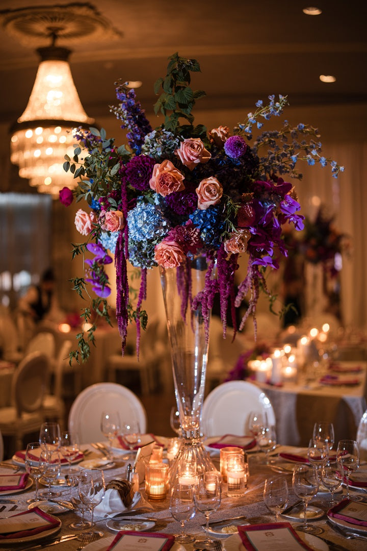 A tall floral arrangement with jewel toned flowers such as roses, hydrangea, orchids, and ameranthus.