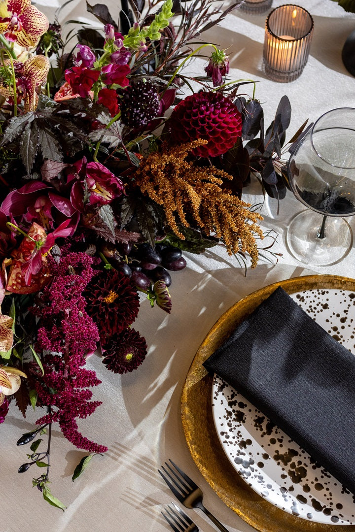 A dark and moody table setting with deep red and purple florals on a table setting of white with black and gold accents.