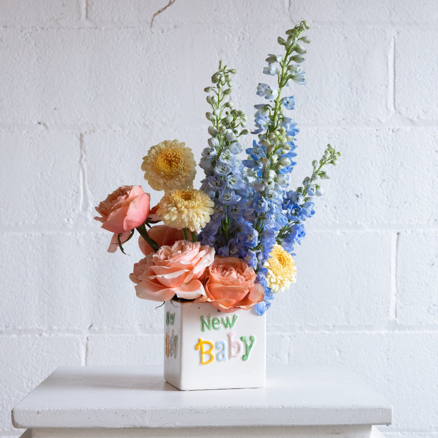 floral arrangement in a container that says 'new baby' with coral, yellow, and blue florals