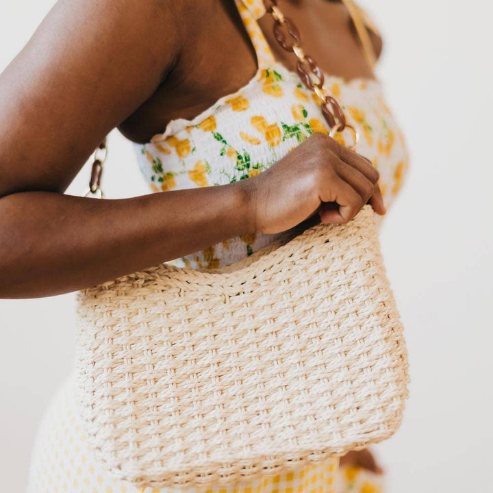 A cream colored basket purse with a brown acrylic chain strap. Model is in a summery dress with lemons on it.