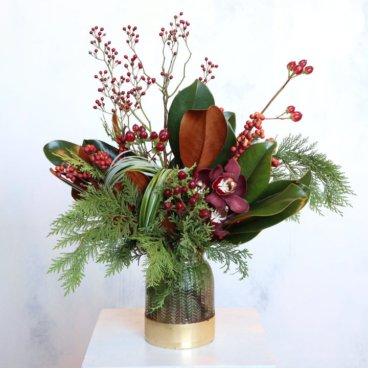 Elegant holiday floral arrangement featuring red berries, magnolia leaves, cedar greenery, and burgundy cymbidium orchids in a gold-accented vase, set against a neutral gray background.