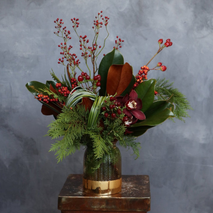 Elegant holiday floral arrangement featuring red berries, magnolia leaves, cedar greenery, and burgundy cymbidium orchids in a gold-accented vase, set against a neutral gray background.