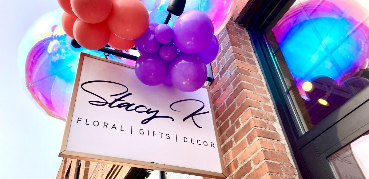 A storefront sign reading 'Stacy K Floral | Gifts | Decor' hangs below a cluster of purple balloons, with large iridescent balloons and a brick building facade in the background.