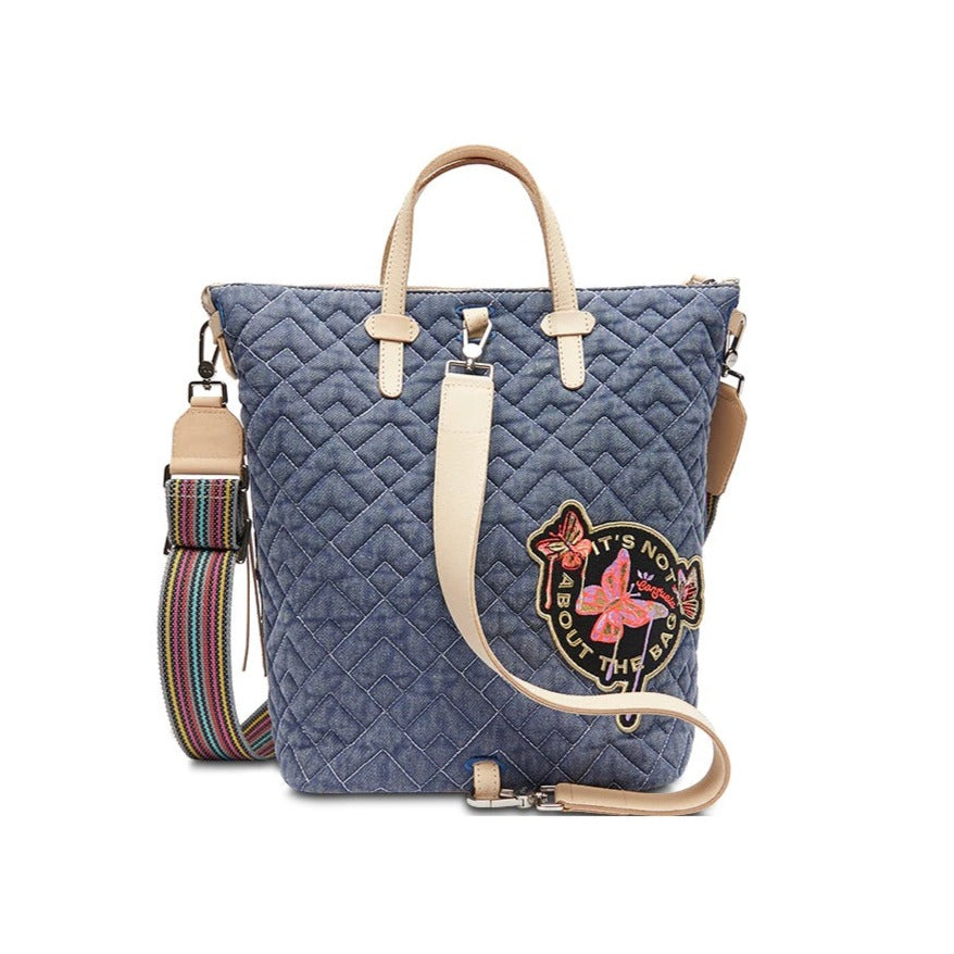 Abby Sling | Consuela | Photo showcasing the reverse side of the bag. Denim blue quilted pattern with deigo leather handles and sling strap. This side of the bag is accented with a colorful butterfly patch that reads "It's Not About The Bag" in gold.