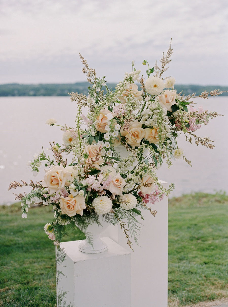 Two floral arrangements on white pedestals. The florals are white, cream, light pink, and green. Some blooms shown are roses, dahlias, and snapdragons.