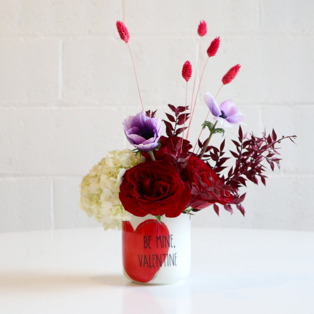 Be Mine Valentine | designed in a keepsake vessel which reads "Be Mine, Valentine". Blooms that  are red roses, purple anemone flowers, white hydrangea, dried bunny tail and red greenery. Vase has a red heart on it. Taken on a white background.
